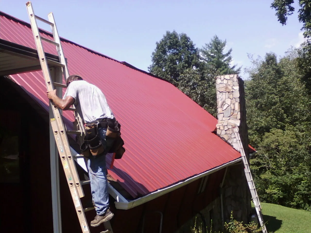 Roofer on a ladder beside a red metal roof and stone chimney — Soffit & Fascia Repair by Roofing Brothers in Free Union